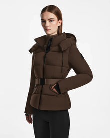 A woman with straight brown hair wears the Rudsak ADELINE - 8124544 Mocha down puffer jacket with a belt and hood, standing against a plain white background and looking forward with a neutral expression.