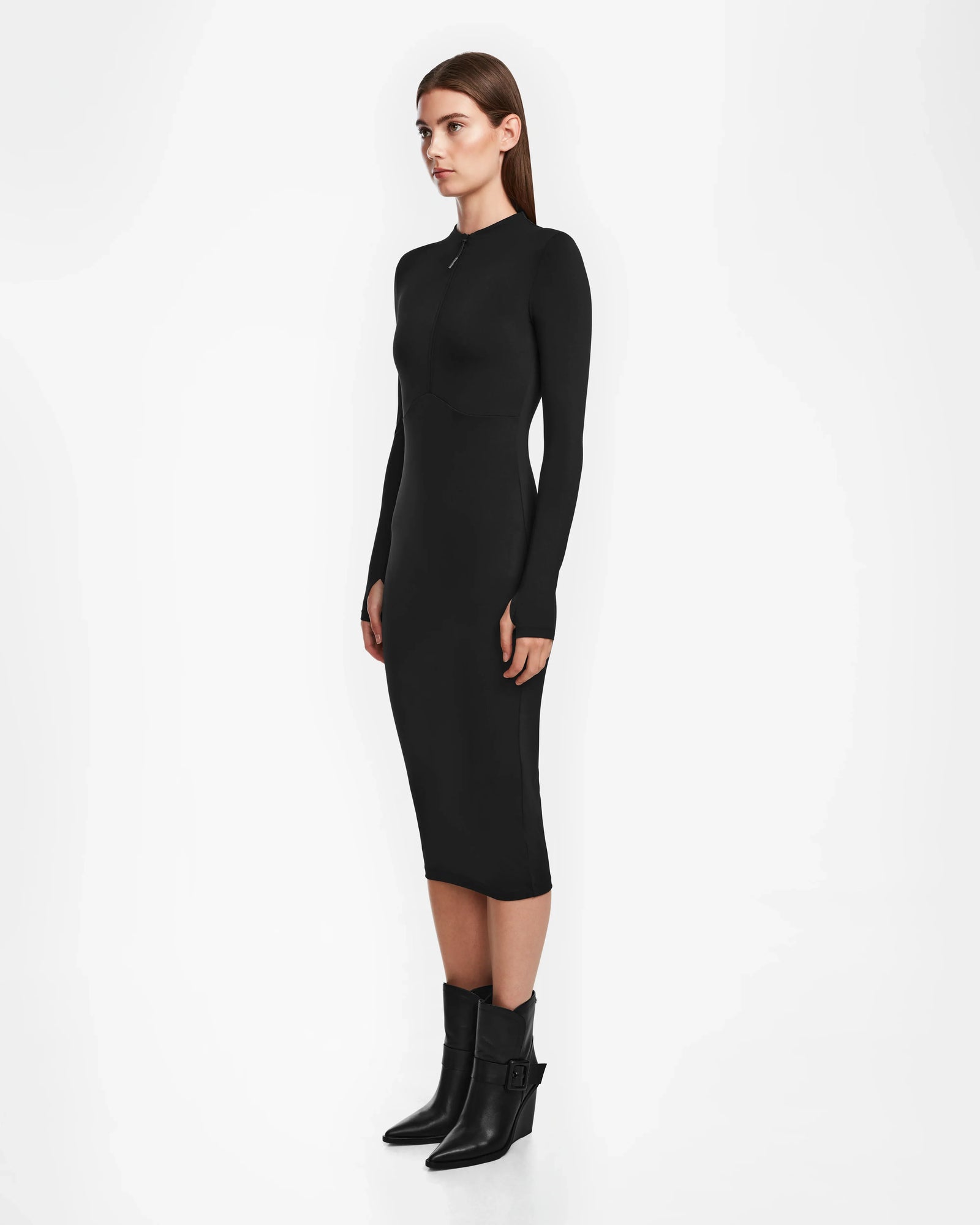A woman stands sideways against a white background, wearing the AGNES - 8524551 Black bodycon stretch dress by Rudsak with black ankle boots. Her straight brown hair is down and she faces forward with a neutral expression.