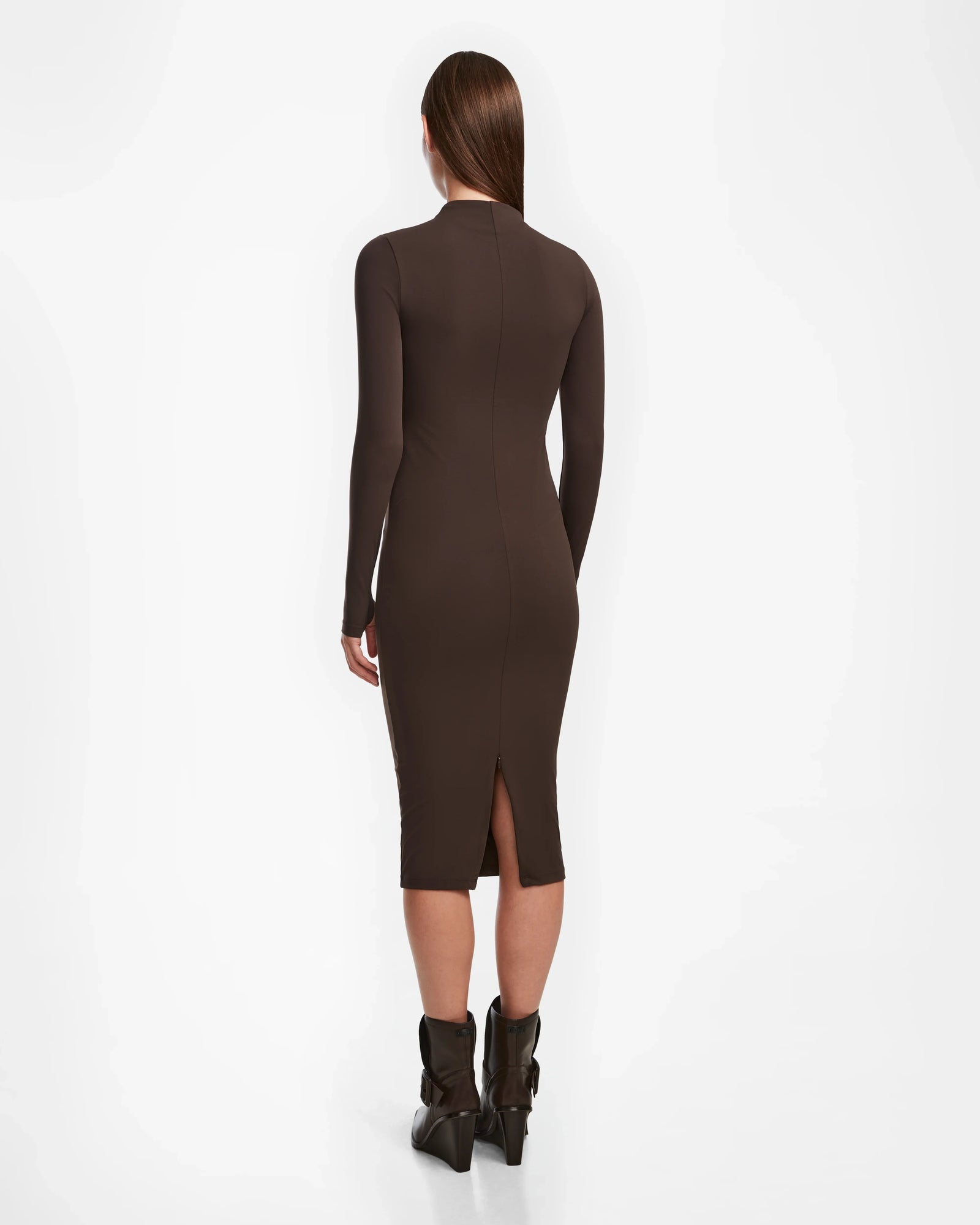 A woman with straight brown hair wears the Rudsak AGNES - 8524551 Espresso fitted, long-sleeve, dark brown bodycon dress and black ankle boots, standing and facing away from the camera against a white background.