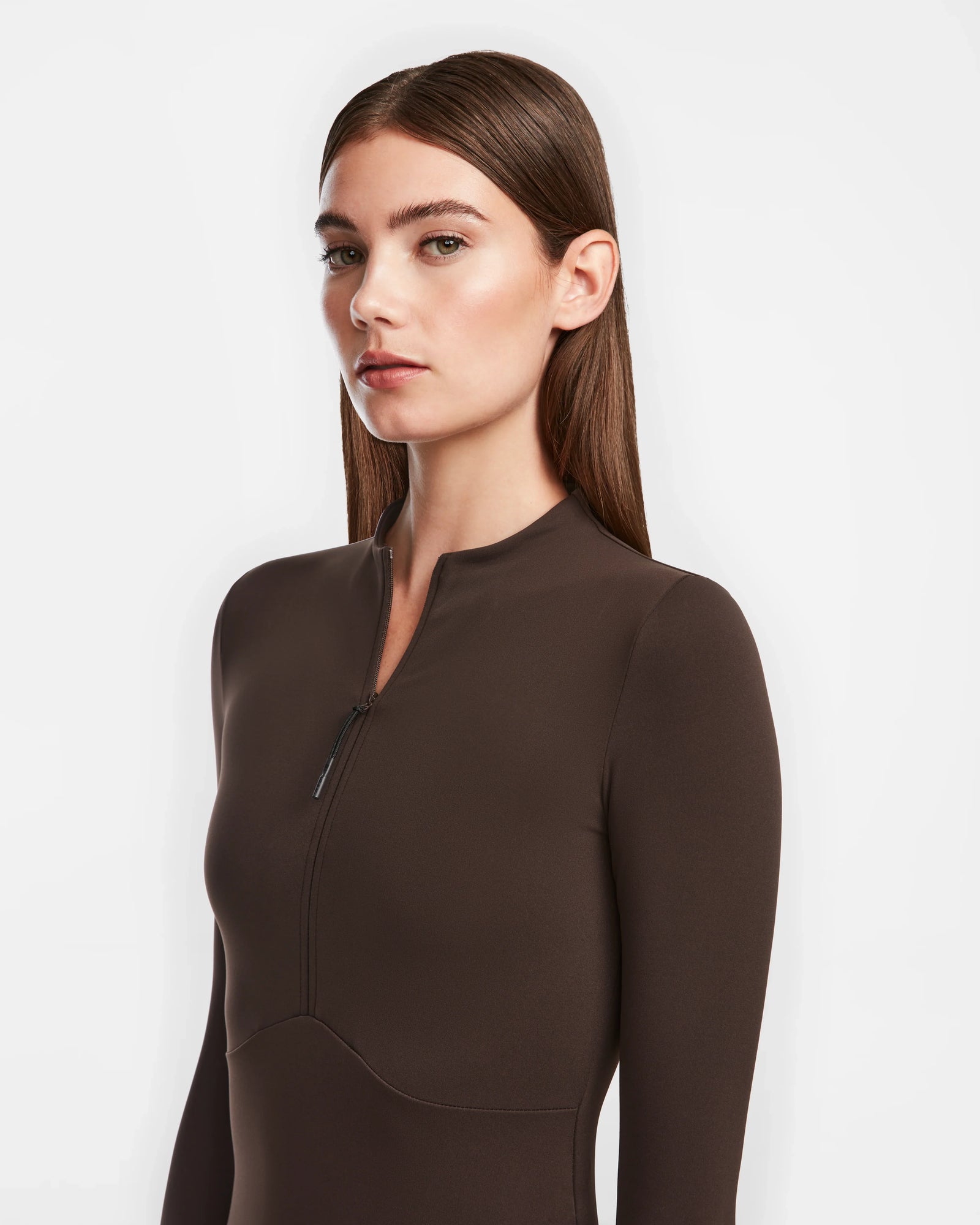 A woman with straight brown hair models the Rudsak AGNES - 8524551 Espresso, a fitted long-sleeve dark brown zip-up dress, standing against a plain white background and gazing slightly to the side.