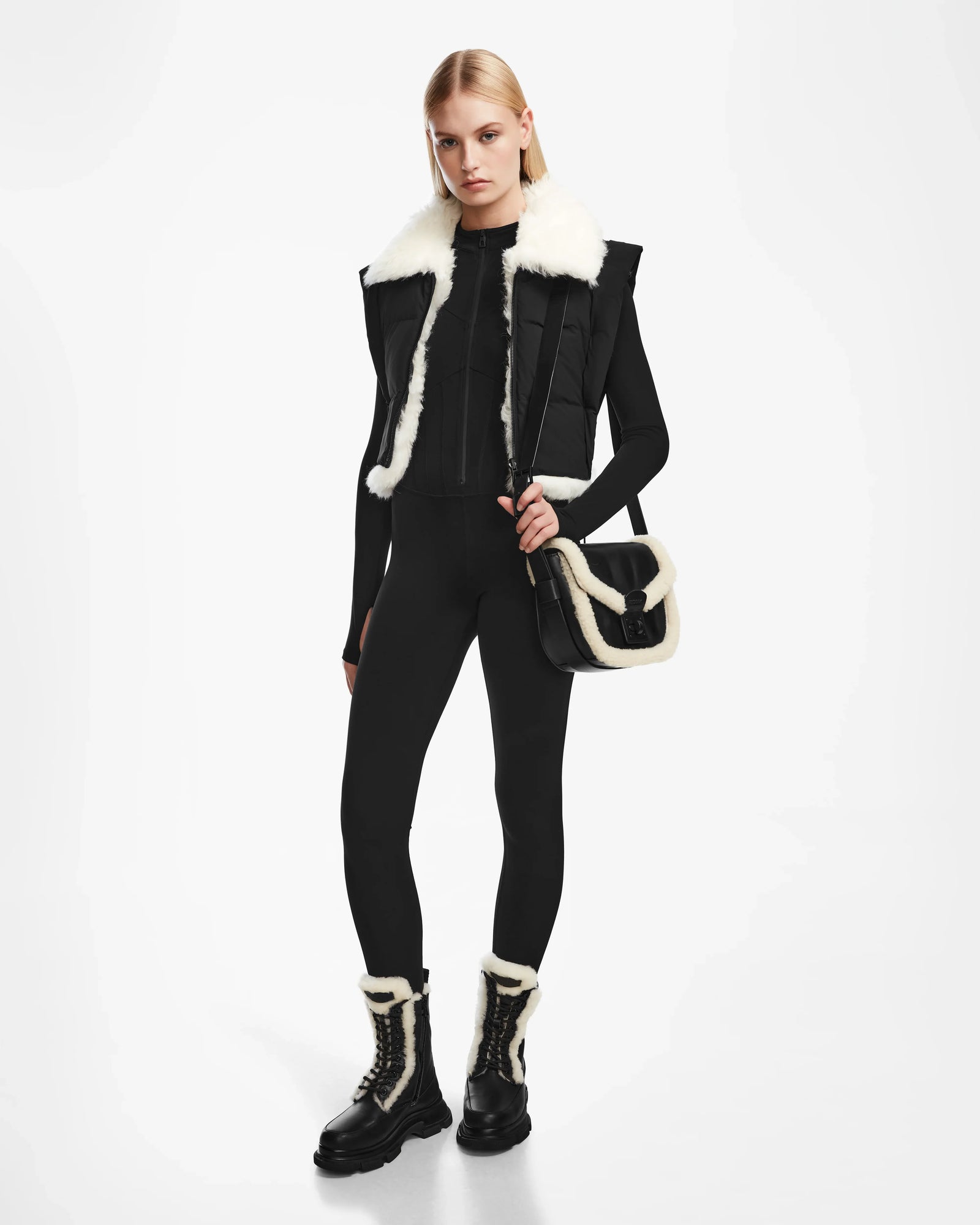 A woman stands against a white background wearing Rudsak’s AIDIA - 8124730 Black fitted outfit, paired with matching fur-trimmed boots, a luxe reversible shearling vest with white faux fur trim, and a black-and-white fur-trimmed shoulder bag.