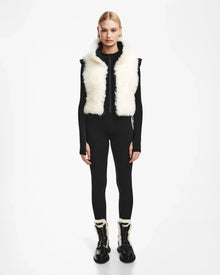 A person stands facing forward, wearing the Rudsak AIDIA - 8124730 Black luxe shearling reversible vest over a fitted black long-sleeve outfit and black boots with white fur trim. The cropped silhouette contrasts with the plain white background.
