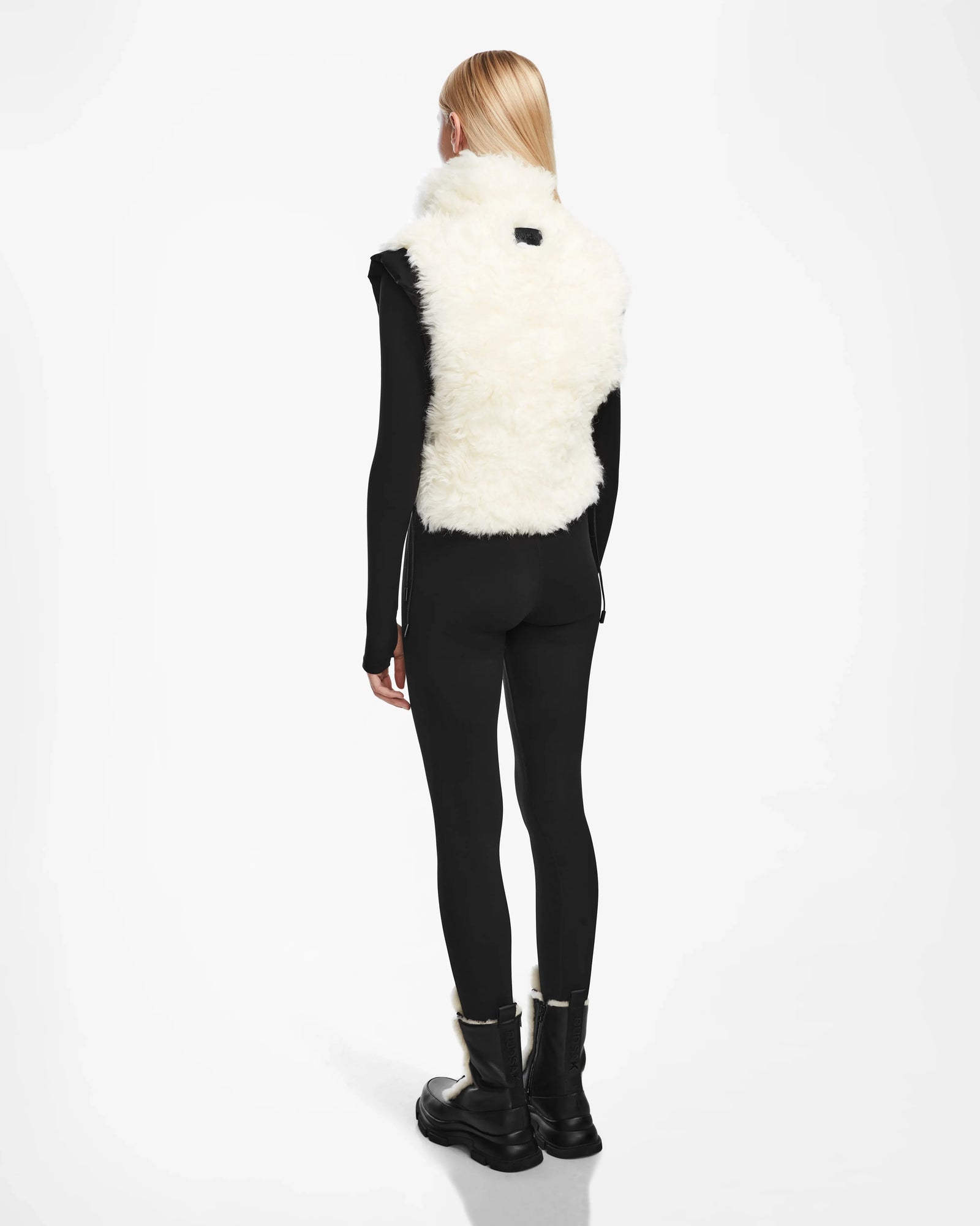 A person with straight blonde hair, wearing black fitted clothes, black boots, and the Rudsak AIDIA - 8124730 Black sleeveless cropped shearling vest, stands facing away against a plain white background.