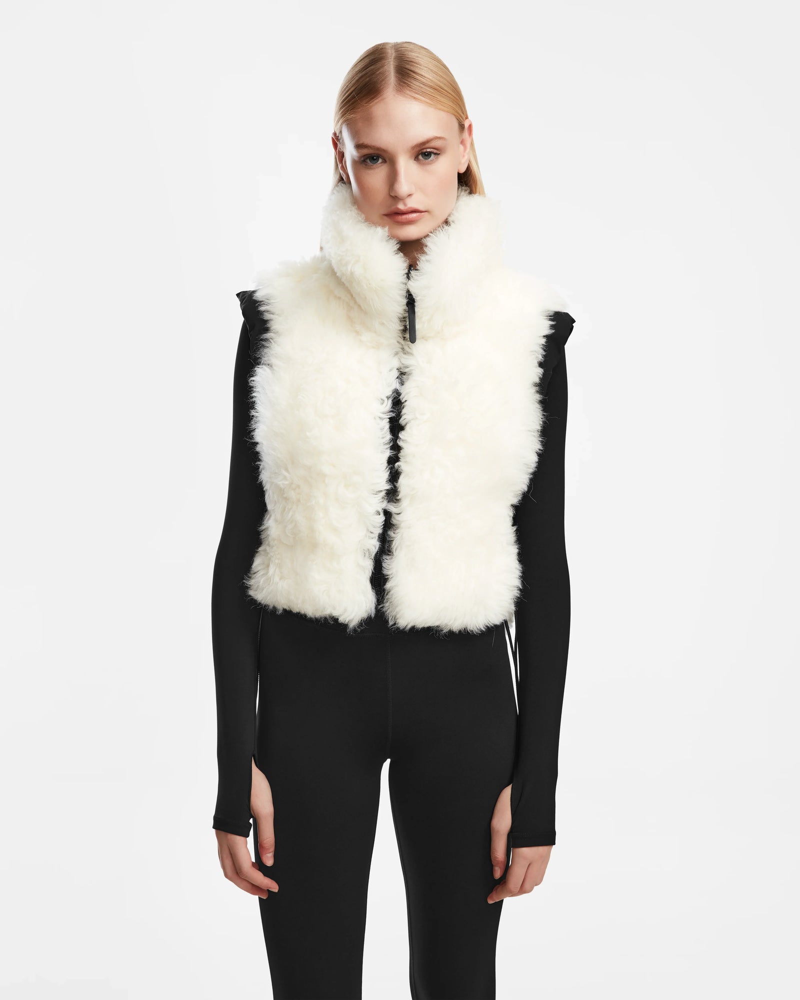 A woman models the Rudsak AIDIA - 8124730 Black, wearing a white shearling cropped vest over a fitted black long-sleeve outfit, standing against a plain white background and facing forward with a neutral expression.