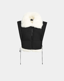 The Rudsak AIDIA - 8124730 Black is a black cropped puffer vest with a large white faux fur collar and hem, front zipper, and side drawstrings, shown on a plain light gray background.