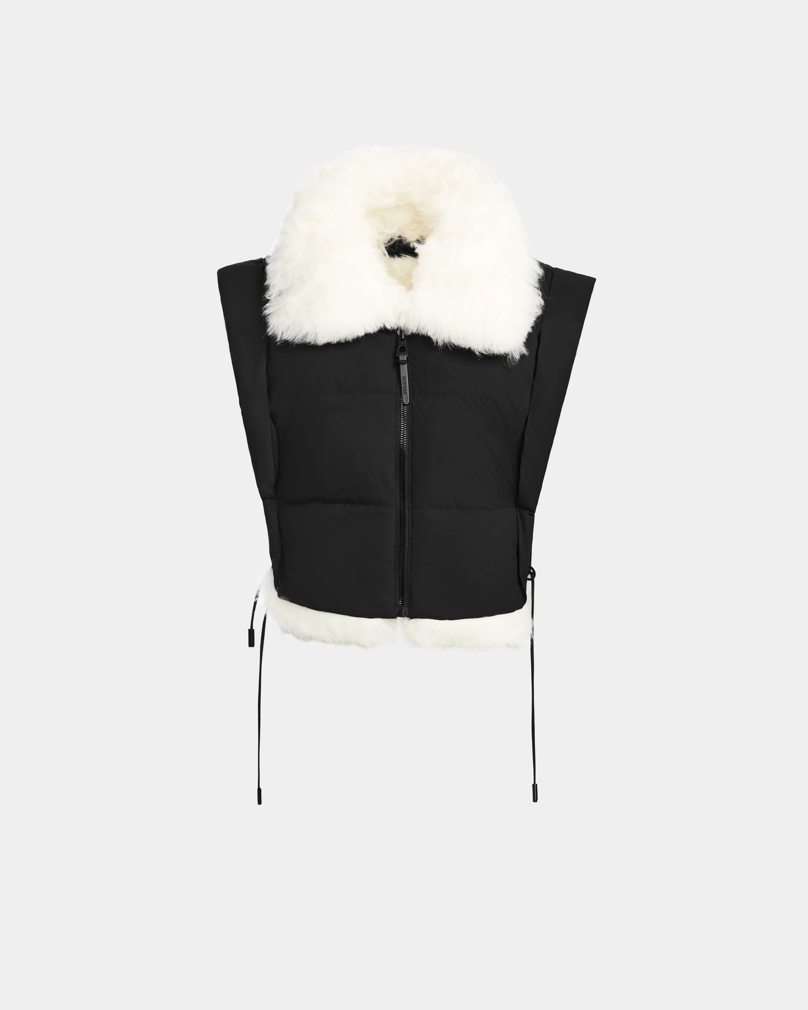The Rudsak AIDIA - 8124730 Black is a black cropped puffer vest with a large white faux fur collar and hem, front zipper, and side drawstrings, shown on a plain light gray background.