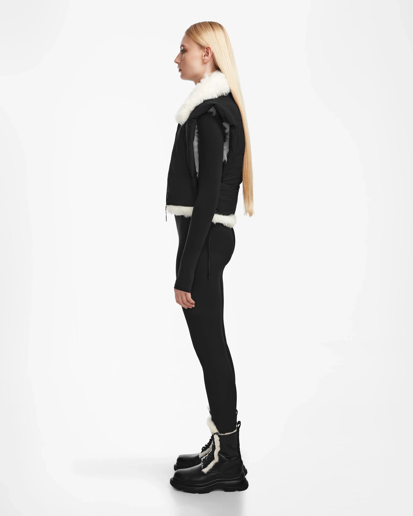 A woman with long blonde hair stands in profile against a plain white background wearing Rudsaks AIDIA - 8124730 Black luxe shearling reversible vest with white fur trim, black leggings, and black winter boots with white laces.
