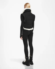 A person with straight blonde hair, wearing the Rudsak AIDIA - 8124730 Black reversible vest with luxe shearling trim, black leggings, and black boots, stands facing away against a plain white background.