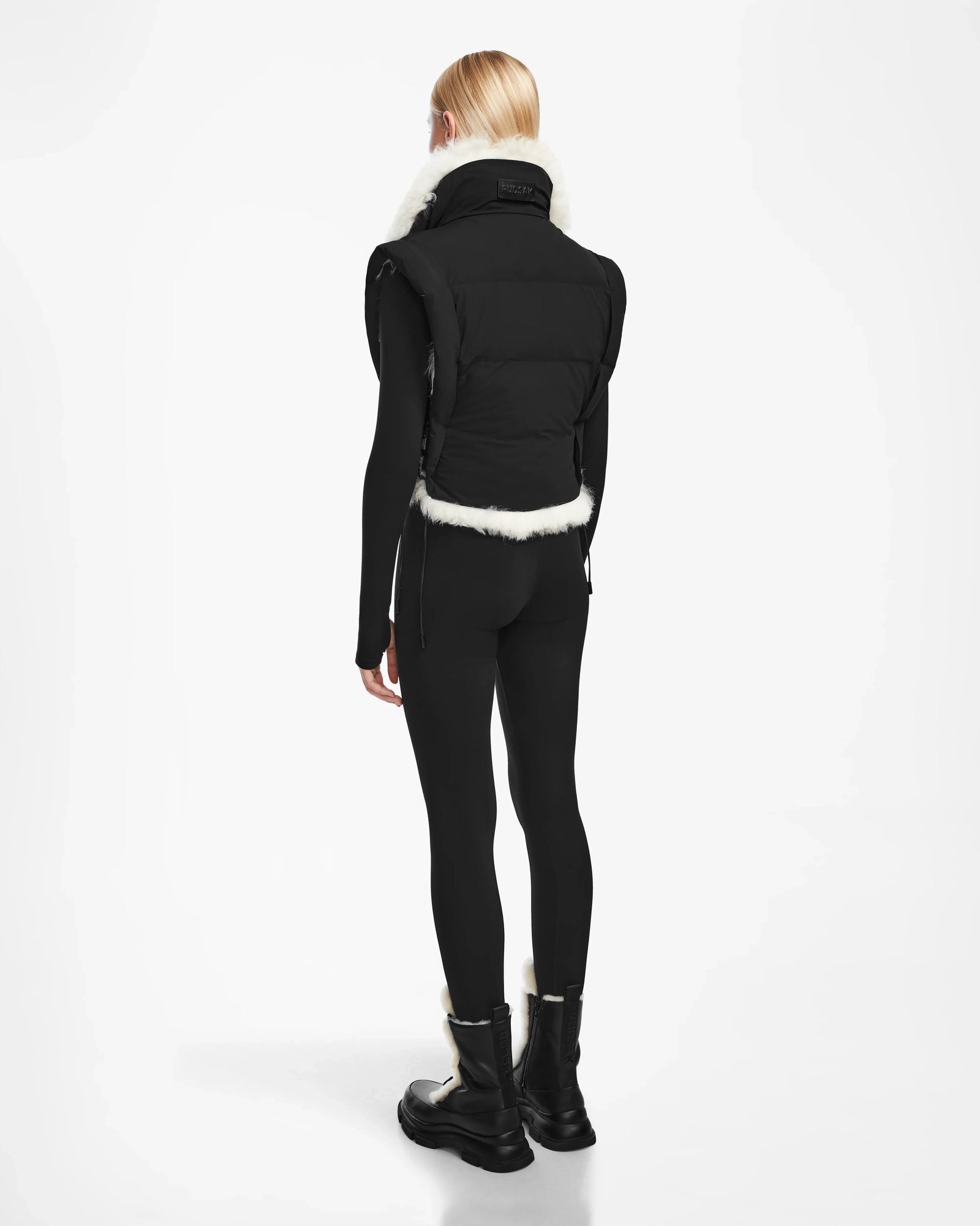 A person with straight blonde hair, wearing the Rudsak AIDIA - 8124730 Black reversible vest with luxe shearling trim, black leggings, and black boots, stands facing away against a plain white background.