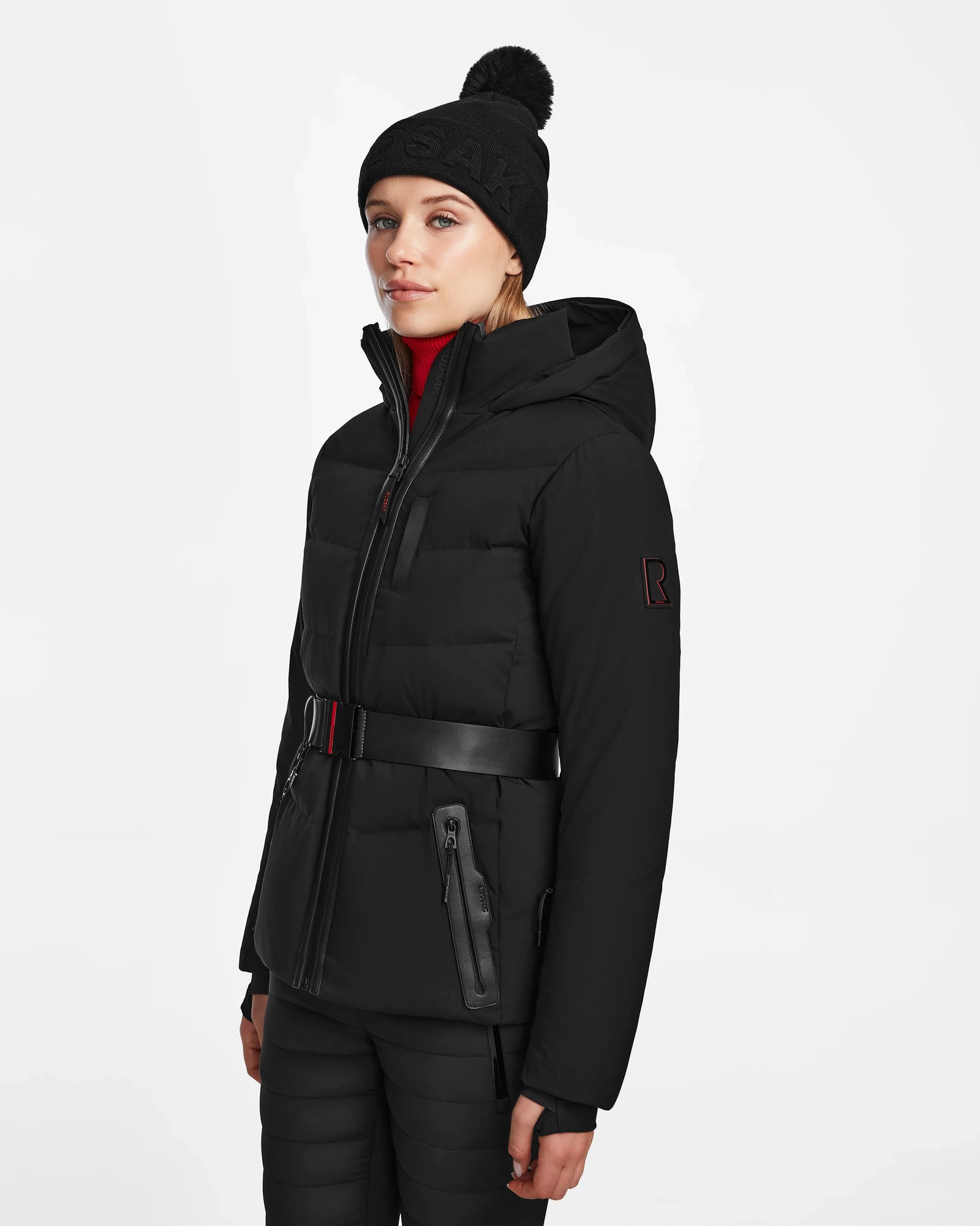 A woman wears the Rudsak ALAIA - 8124816 black quilted ski jacket with a belt, paired with black pants and a pom-pom beanie, standing against a plain white background.