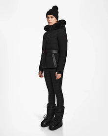 A woman wears the Rudsak ALAIA - 8124816 Black womens quilted ski jacket with a fur-trimmed hood, belted puffer design, matching pants, black beanie, and winter boots—ideal for the slopes—against a white background.