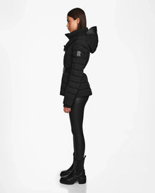 A woman stands in profile against a white background, wearing the Rudsak ALISA - L124506 Black quilted down puffer jacket, black leggings, and black chunky boots.
