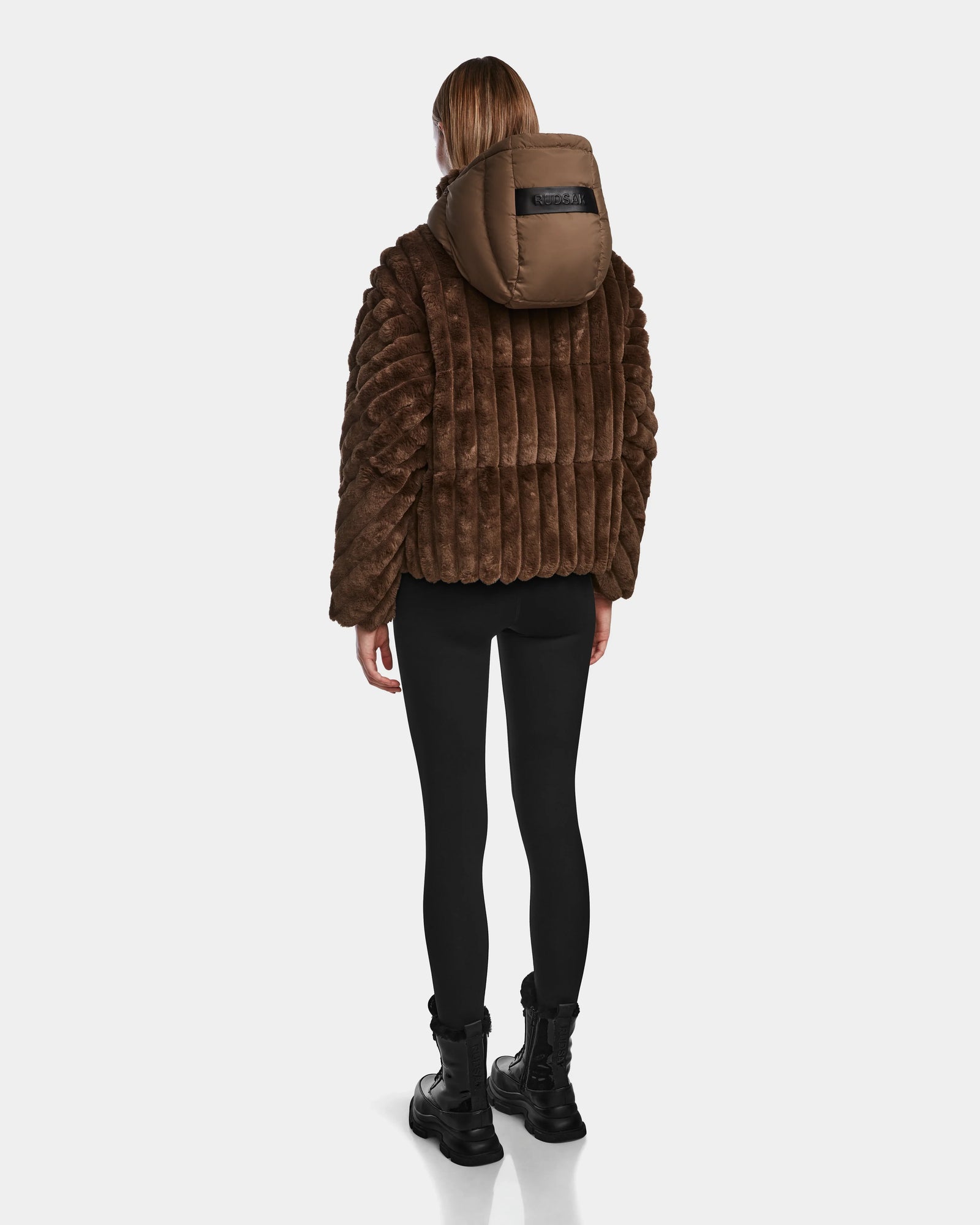 A person stands facing away, wearing the Rudsak ANASTASIA - 8124804 Espresso brown velvet puffer jacket with a hood, paired with black leggings and black boots, against a plain white background.
