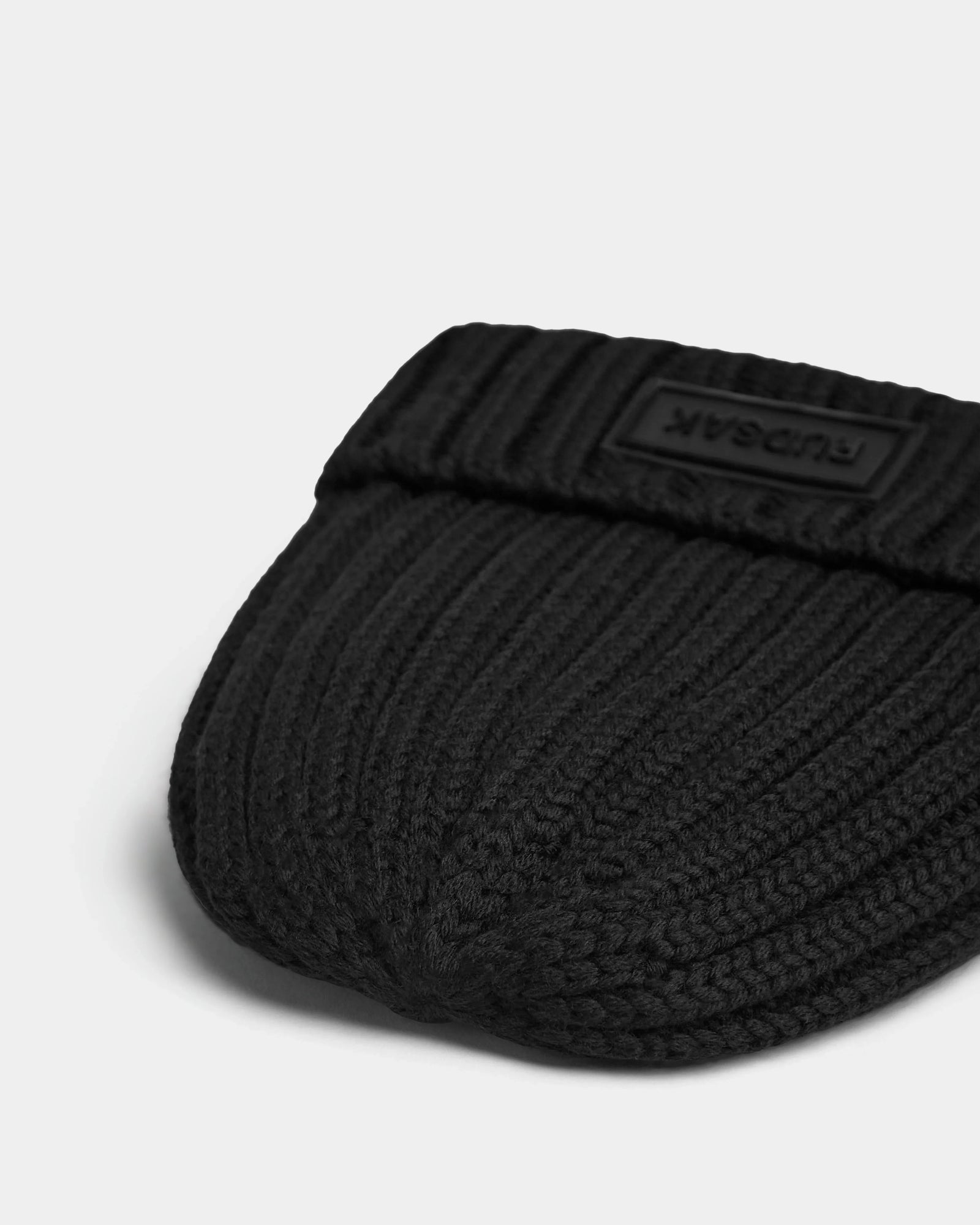 A close-up of the ANISA - L423500 Black/Black ribbed knit beanie by Rudsak, featuring a folded brim and a black patch with raised branding. The hat rests on a plain white surface—an essential for any winter wardrobe.