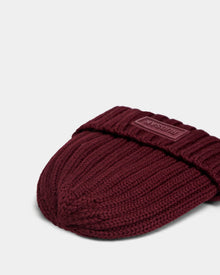 A close-up of the ANISA - L423500 Wine ribbed knit beanie by Rudsak with a folded cuff and rectangular patch. This cozy winter accessory pops against a plain light gray background, perfect for updating your cold-weather wardrobe.