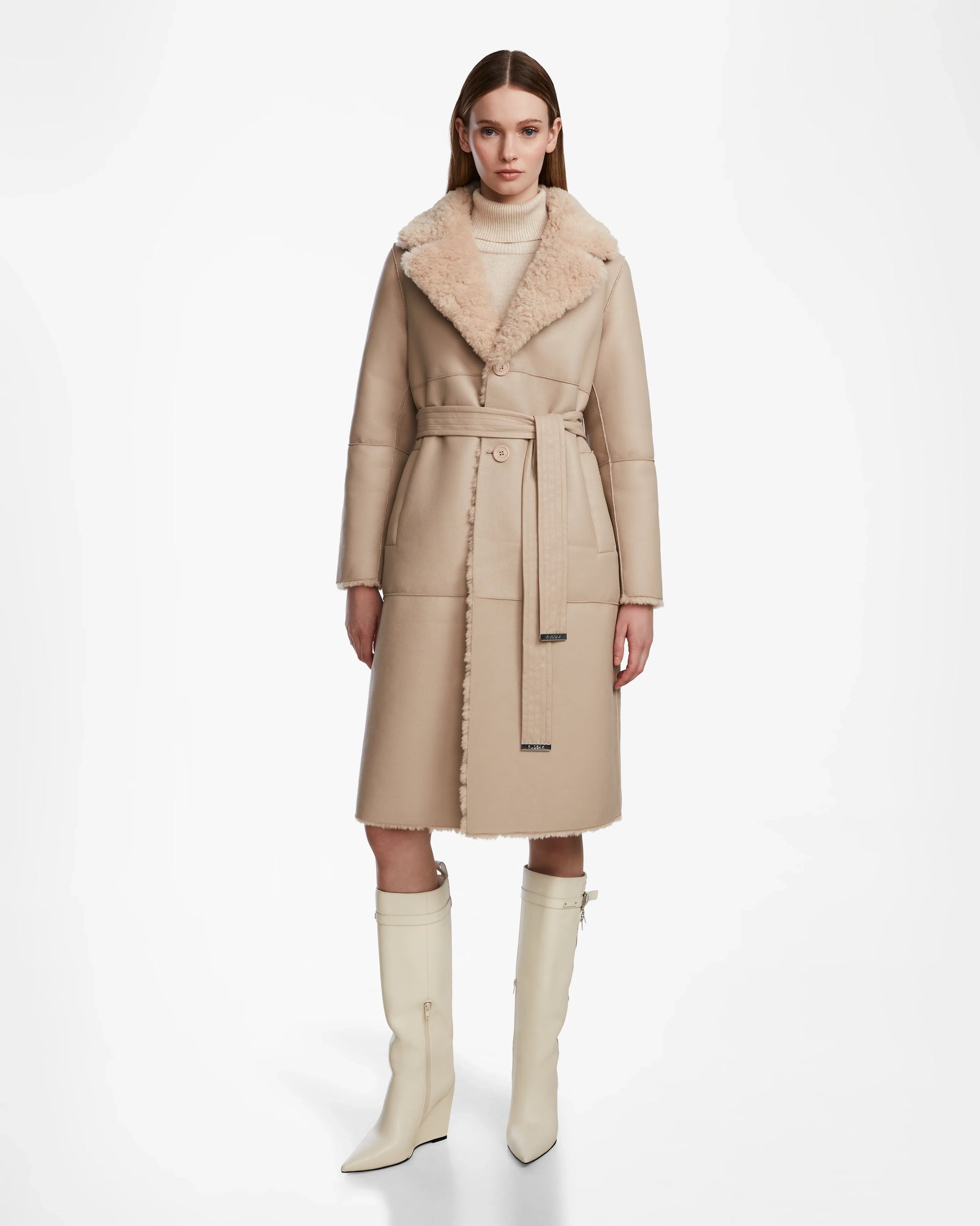 A woman faces forward in the ANNABELLE - L124544 Sand vegan leather coat by Rudsak, featuring a belted waist and shearling collar. She pairs it with a light turtleneck and cream knee-high boots against a plain white background.