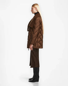 A woman with long blonde hair stands in profile, wearing the Rudsak ANNE MARIE - 8124713 Mocha quilted down wool jacket with a matching scarf, dark brown skirt, and black knee-high boots against a plain white background.