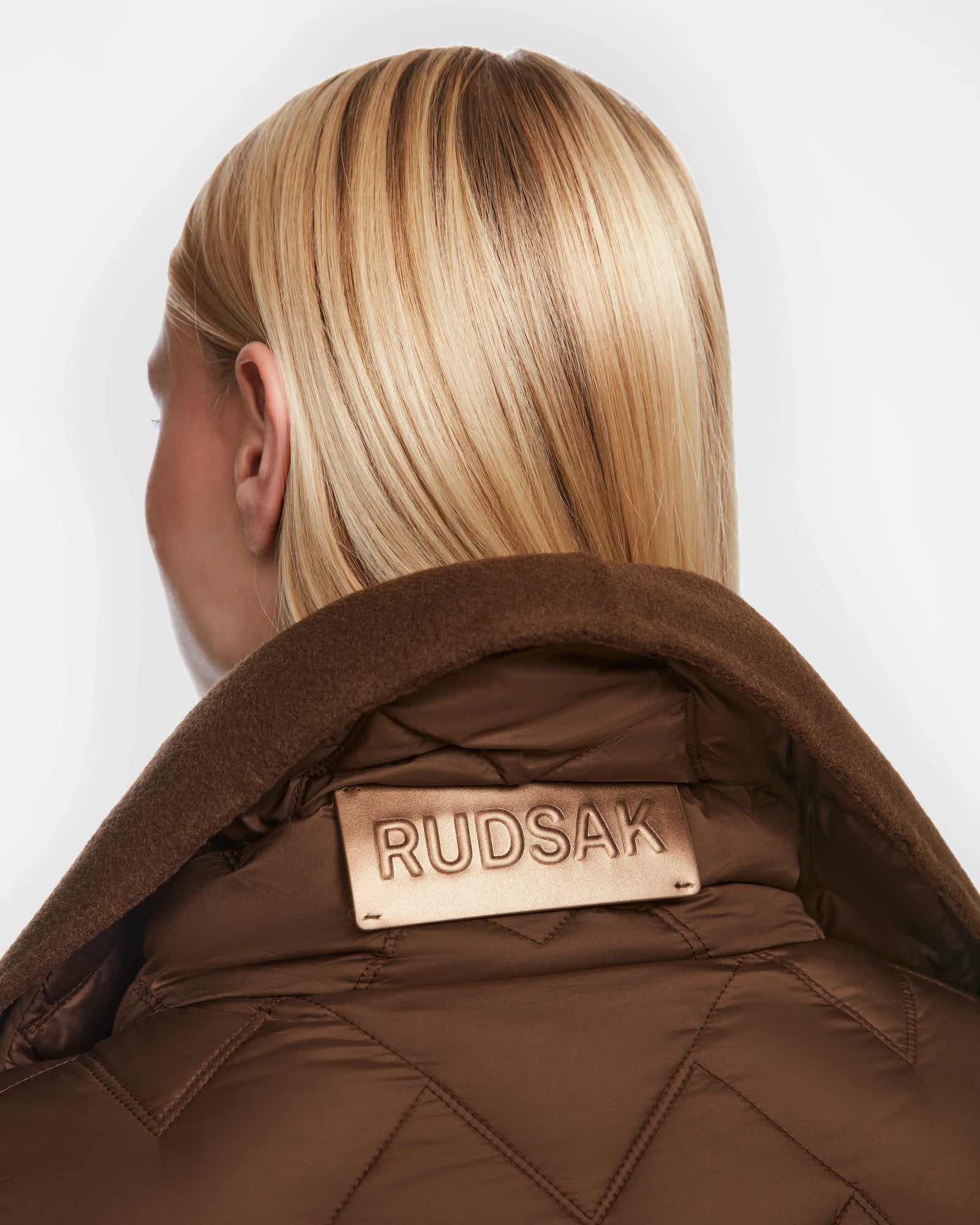 A person with straight blonde hair is shown from the back, wearing the Rudsak ANNE MARIE - 8124713 jacket in mocha quilted fabric with a large collar, featuring a beige Rudsak label prominently displayed on the collar.