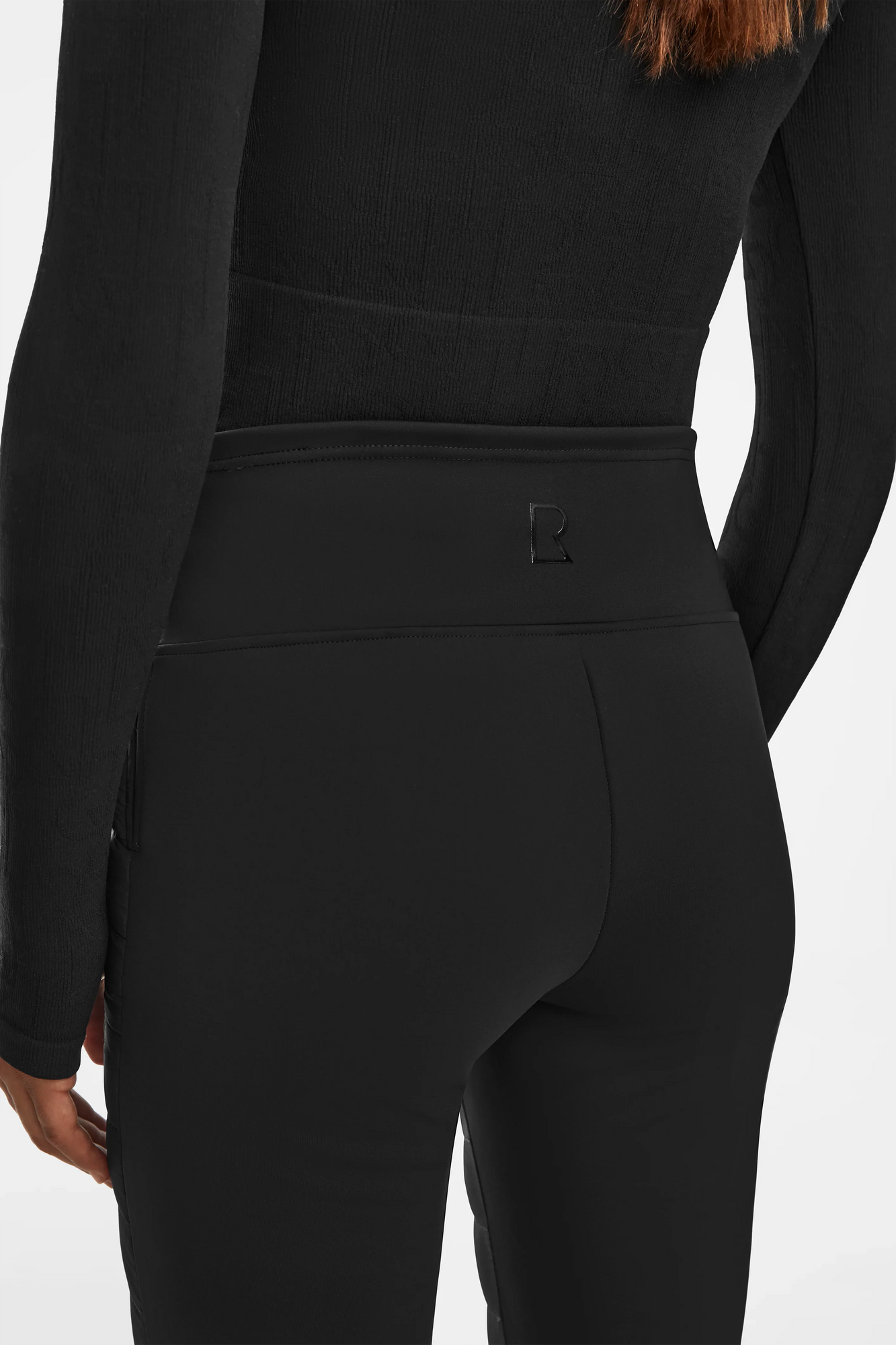 A person wears a fitted black long-sleeve top and high-waisted Rudsak ARIL M - 8522514 BLACK sustainable insulated pants, shown from the back. The pants have a small stitched logo on the waistband for an après-ski chic look.