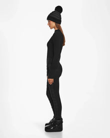 Woman in side profile wears Rudsak ARIL M - 8522514 BLACK beanie with pom-pom, a black long-sleeve top, après-ski chic womens quilted pants, and black platform boots, standing against a white background.