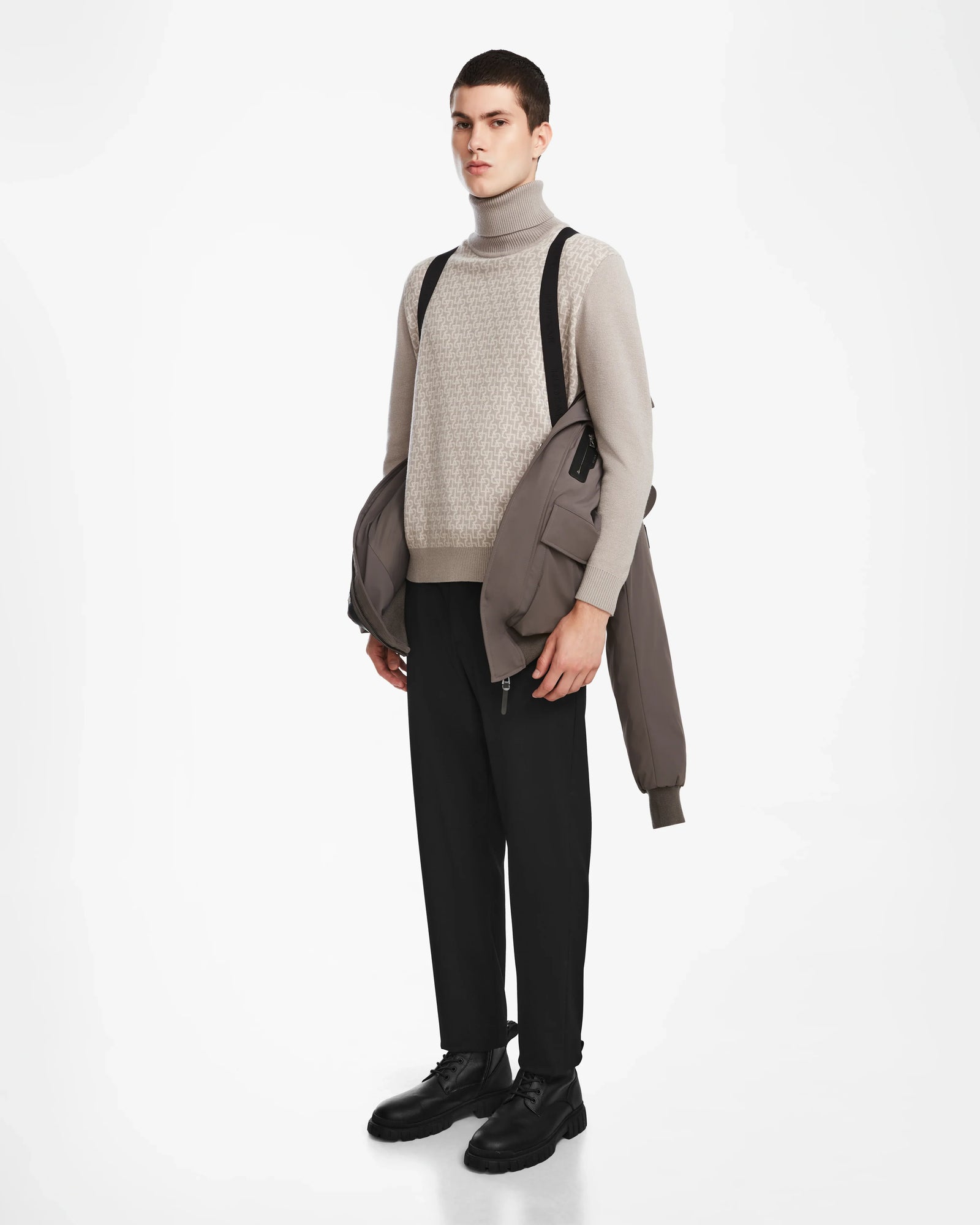 A person stands against a white background wearing Rudsaks ASH - 6124599 Grey water-resistant bomber winter jacket draped over one arm, a beige turtleneck, black pants and shoes, with black backpack straps on their shoulders.