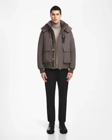 A man faces forward wearing the Rudsak ASH - 6124599 Grey water-resistant bomber over a beige turtleneck, black pants, and black shoes, set against a plain white background.