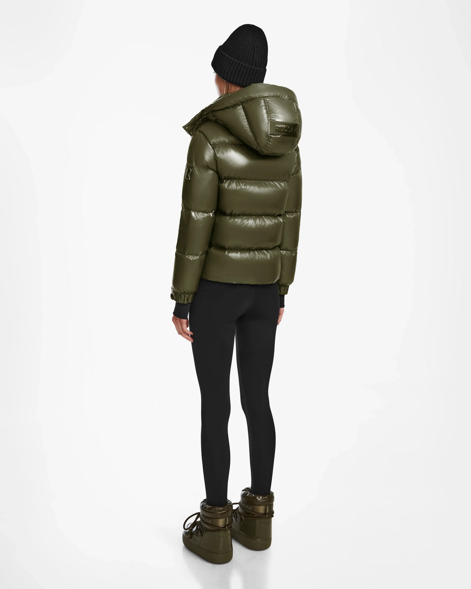 A person in the Rudsak ASHLYN S - 8124565 Loden Green puffer jacket, black leggings, chunky olive boots, and a black beanie stands facing away against a plain white background.