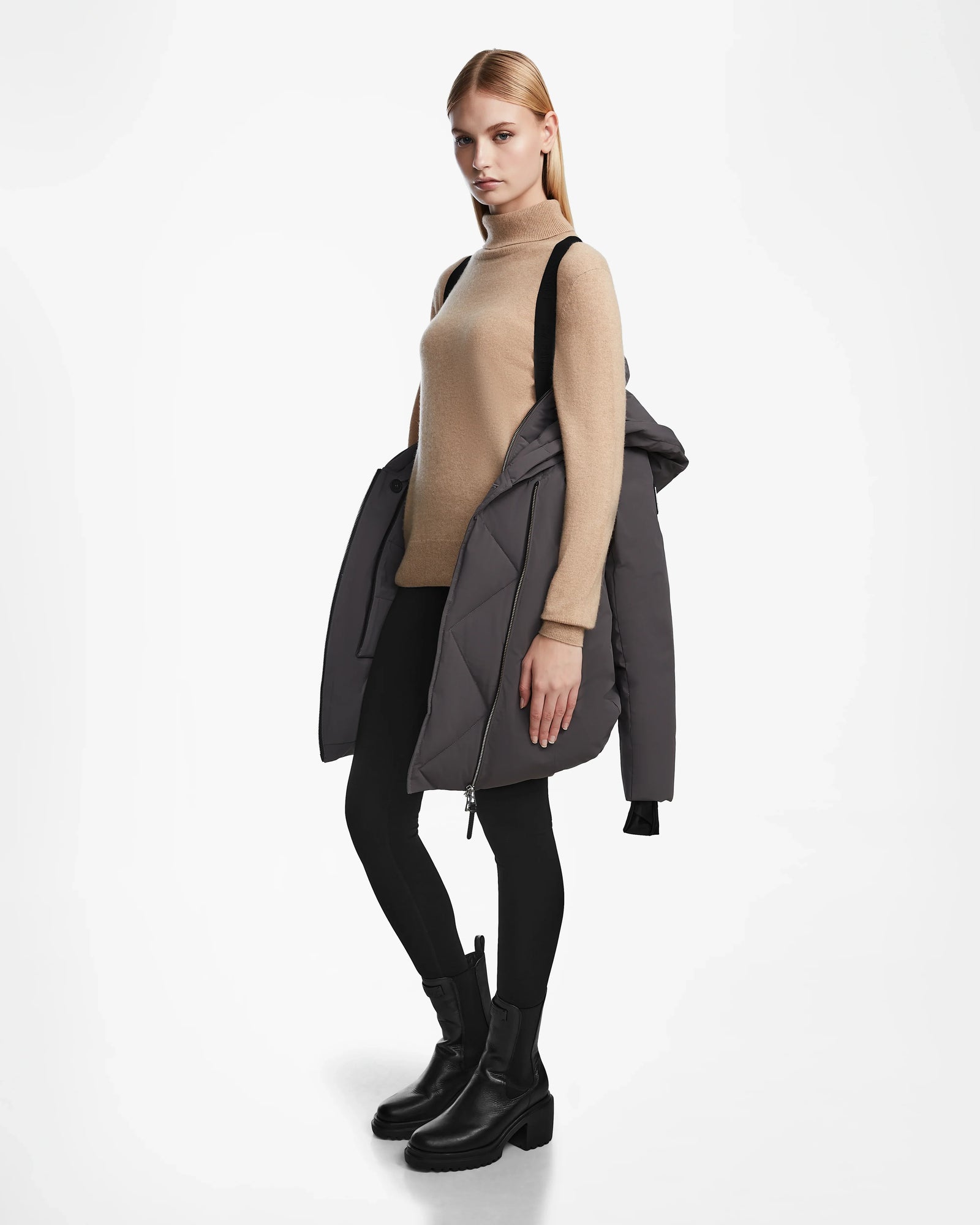 A woman with straight blonde hair wears a beige turtleneck, black leggings, and ankle boots. She carries the Rudsak AURORE - L123561 Grey winter coat over one shoulder, standing against a plain white background.