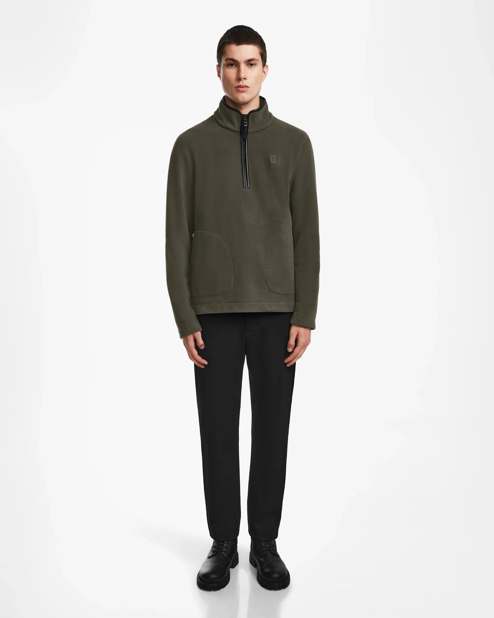 A person stands facing forward, wearing the Rudsak BECK - 6524538 Army mens fleece pullover with vegan leather trims, black pants, and black shoes against a plain white background.