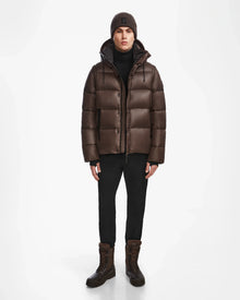 A person wearing the Rudsak BOBBY - 6124506 Espresso men’s quilted down puffer jacket, brown knit beanie, black pants, and brown lace-up boots stands against a plain white background.