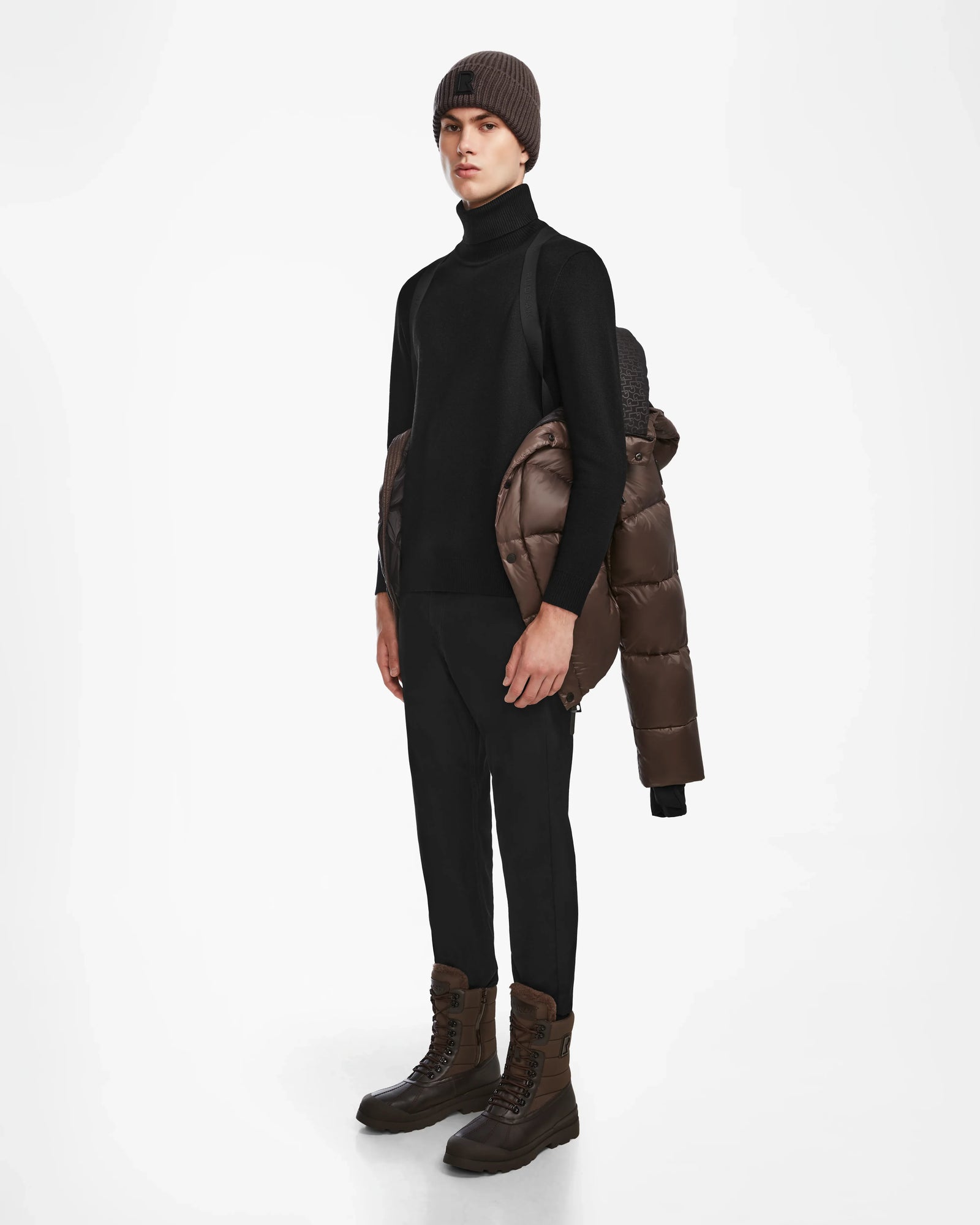 A person stands against a white background in black clothes and brown boots, holding the Rudsak BOBBY - 6124506 Espresso mens quilted down puffer with a water-resistant shell draped over one shoulder.