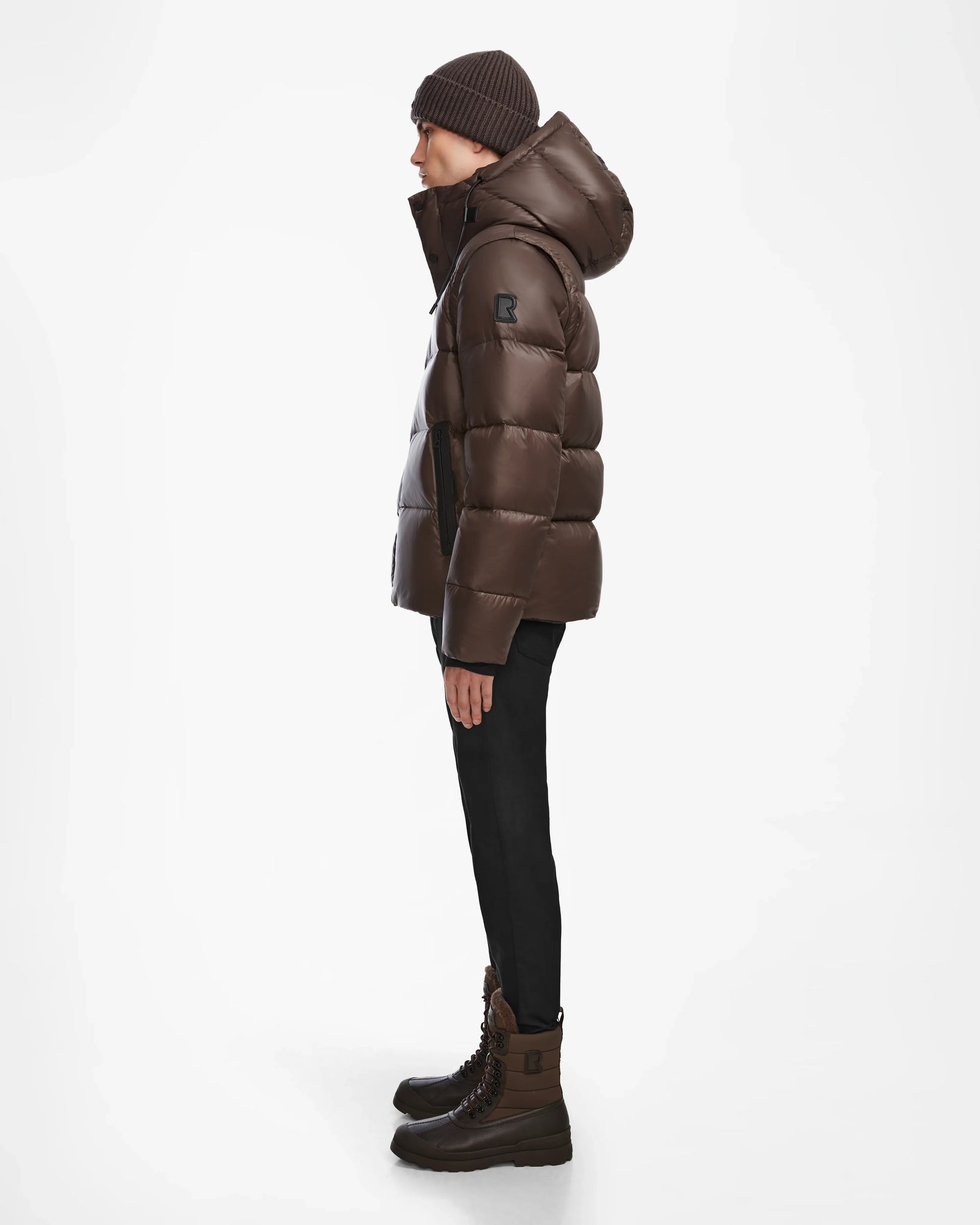 A person in a Rudsak BOBBY - 6124506 Espresso mens quilted down puffer with a water-resistant shell, brown winter boots, black pants, and a brown knit beanie stands in profile against a plain white background.