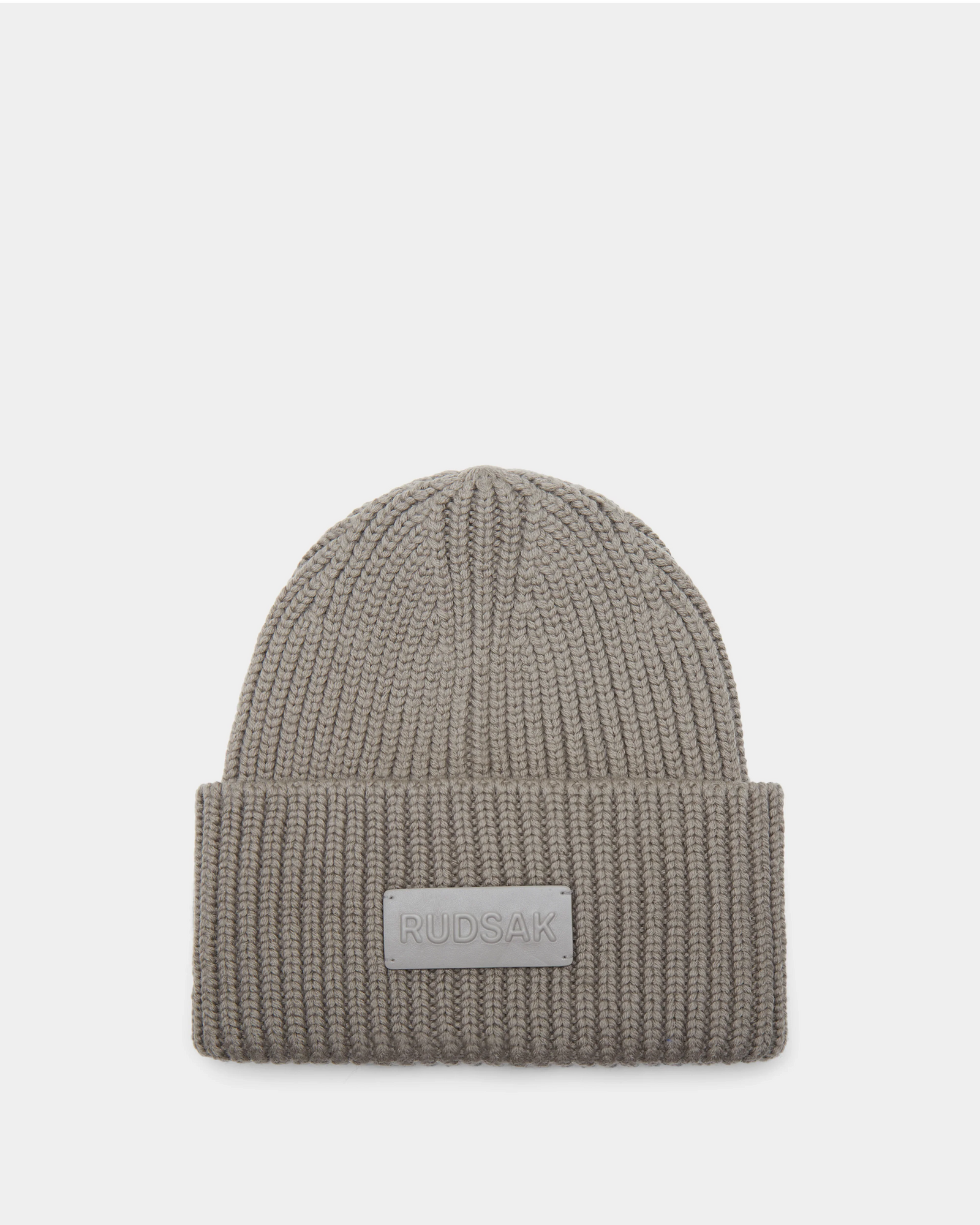 The Rudsak COLIN - 9424518 Cement is a taupe unisex ribbed beanie with a thick folded brim and a rectangular Rudsak patch on the front, shown against a plain white background.