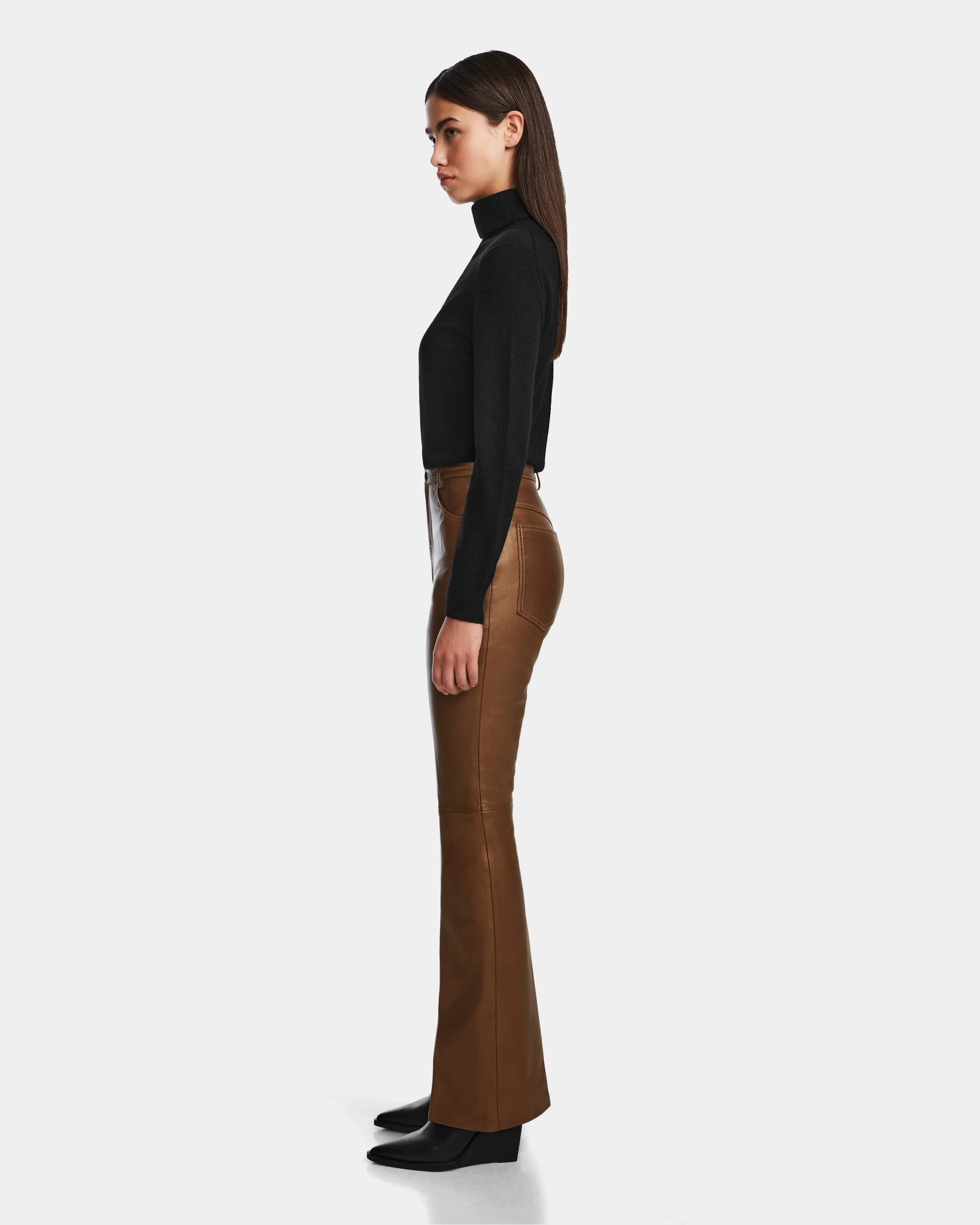 A woman stands in profile against a plain background, wearing a black long-sleeve turtleneck top paired with Rudsak’s CARISSA - 8524545 Antique Bronze leather pants and black boots.