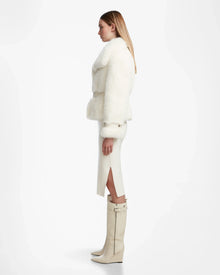 A woman in profile stands against a white background wearing the Rudsak CHLOE - L124564 Iceland faux fur jacket with a white midi skirt and knee-high heeled boots, her sleek blond hair down—an elegant winter look.