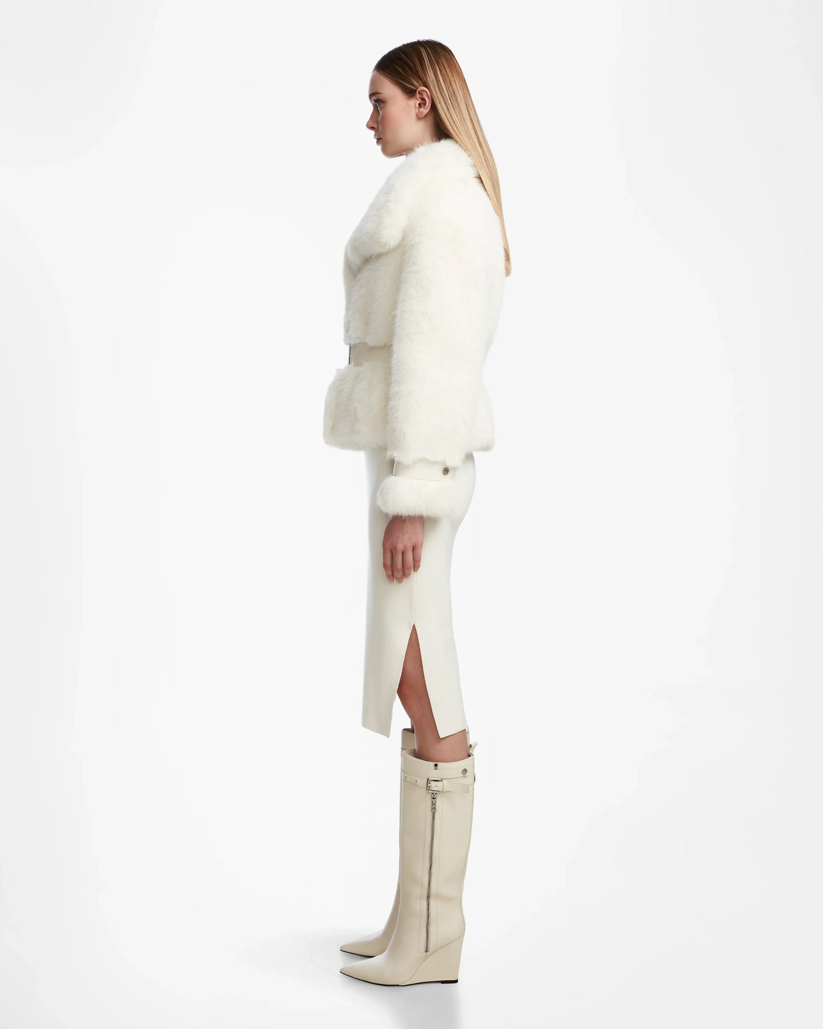 A woman in profile stands against a white background wearing the Rudsak CHLOE - L124564 Iceland faux fur jacket with a white midi skirt and knee-high heeled boots, her sleek blond hair down—an elegant winter look.