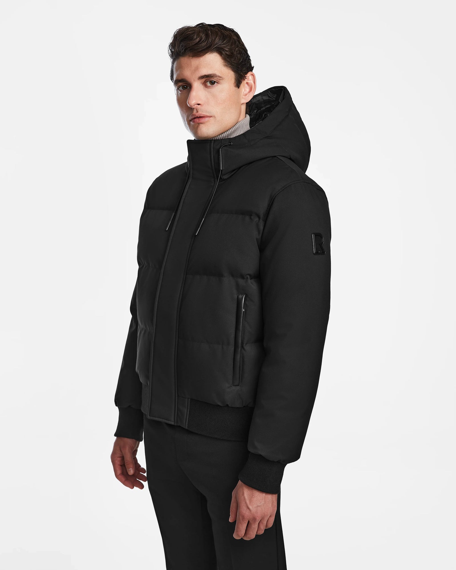 A man models the Rudsak DALLAS - M122501 Black weather-resistant jacket and black pants, standing against a plain white background and gazing slightly to the side with a neutral expression.