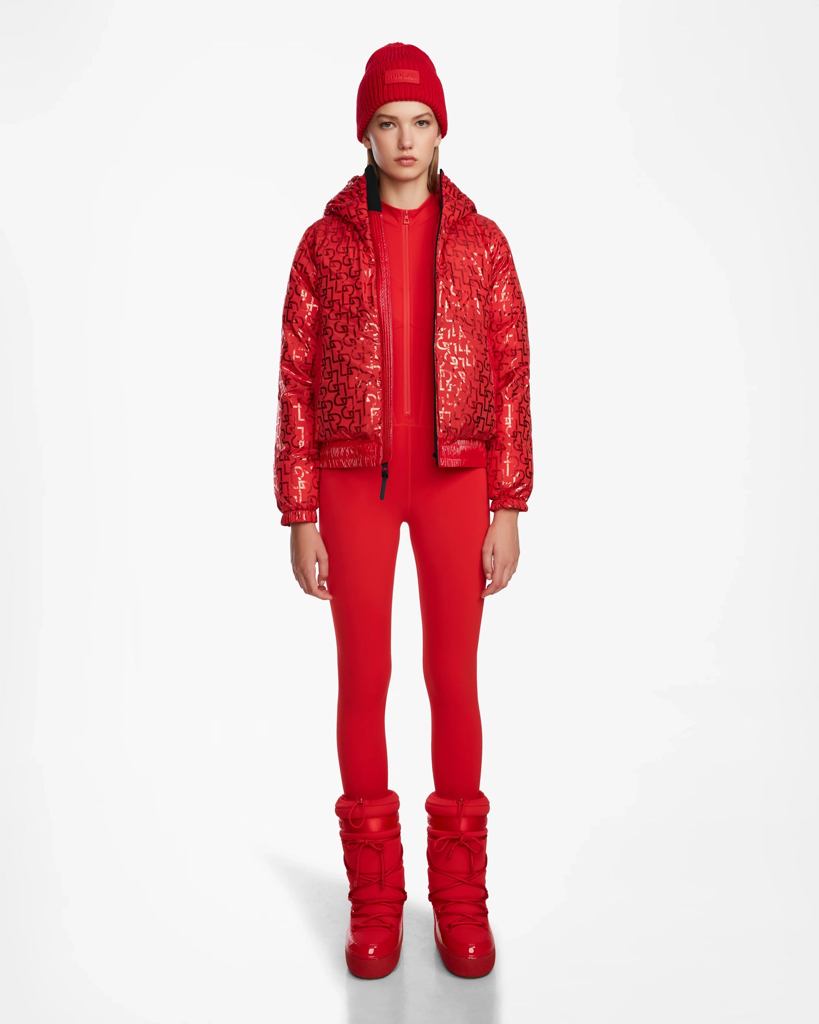 A person poses against a white background wearing the Rudsak DANI M - 8124539 Flame Red shiny bomber-style ski jacket, paired with a red beanie, red leggings, and red snow boots, sporting a neutral expression.