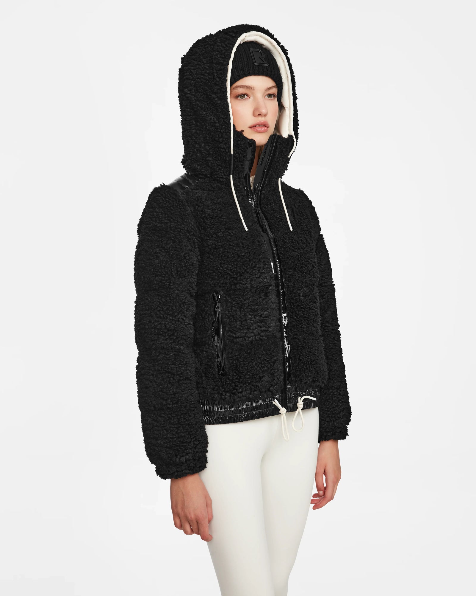A person wears the Rudsak DANI S - 8124537 Black bomber-style ski jacket with cream drawstrings, white leggings, and a black beanie, standing against a plain white background.