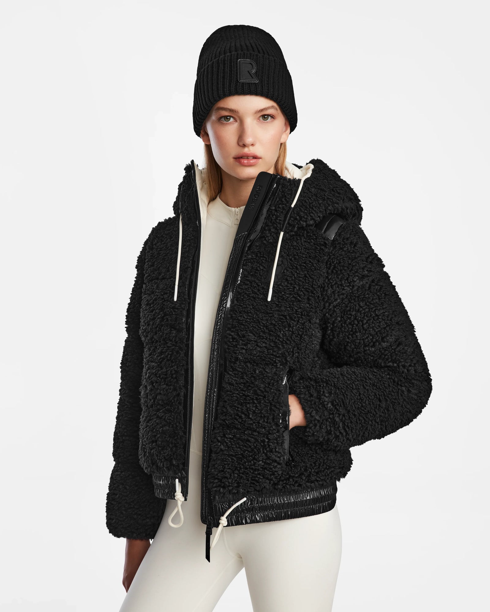 A woman wears the Rudsak DANI S - 8124537 Black bomber-style ski jacket with hood, a black beanie, and cream pants, standing facing forward against a plain white background.