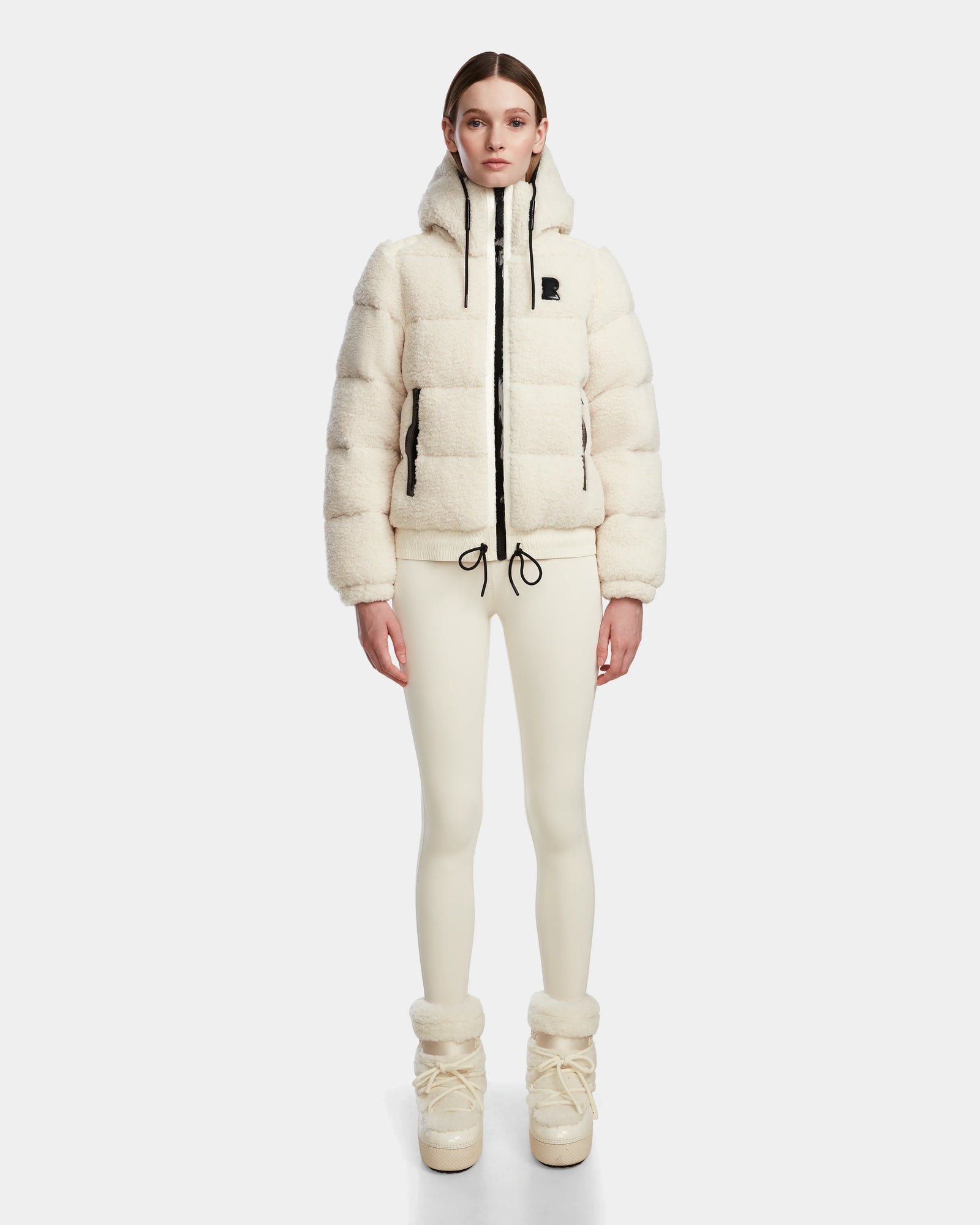 A woman faces forward in a minimalist white studio, wearing the cream-colored Rudsak DANI S - 8124537 Iceland cropped ski jacket with hood, paired with cream leggings and matching boots.