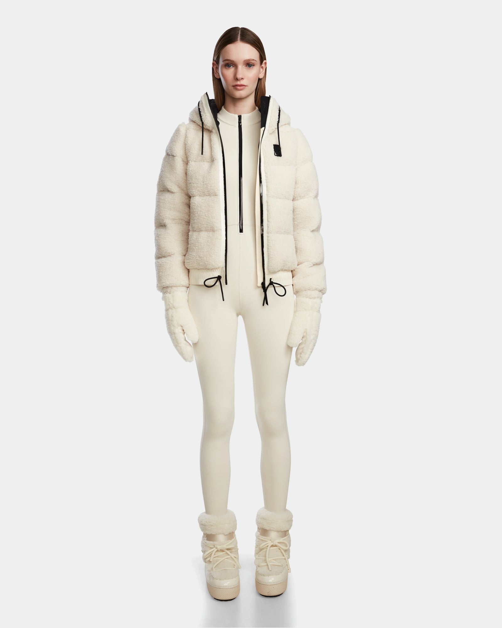 A person stands facing forward in Rudsak’s DANI S - 8124537 Iceland: an all-cream ensemble with a hooded cropped ski jacket, fitted bodysuit, gloves, and fur-detailed boots. The plain white background highlights this winter look.
