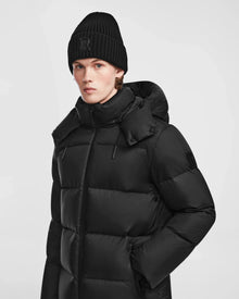 A person wearing the Rudsak DANIEL M - M124510 Black mens hooded down puffer and a black knit beanie stands against a plain light background, looking directly at the camera with a neutral expression.