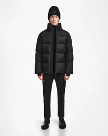 A person stands facing forward against a plain white background, wearing the Rudsak DANIEL M - M124510 Black weather-resistant puffer jacket with black pants, boots, and a beanie.