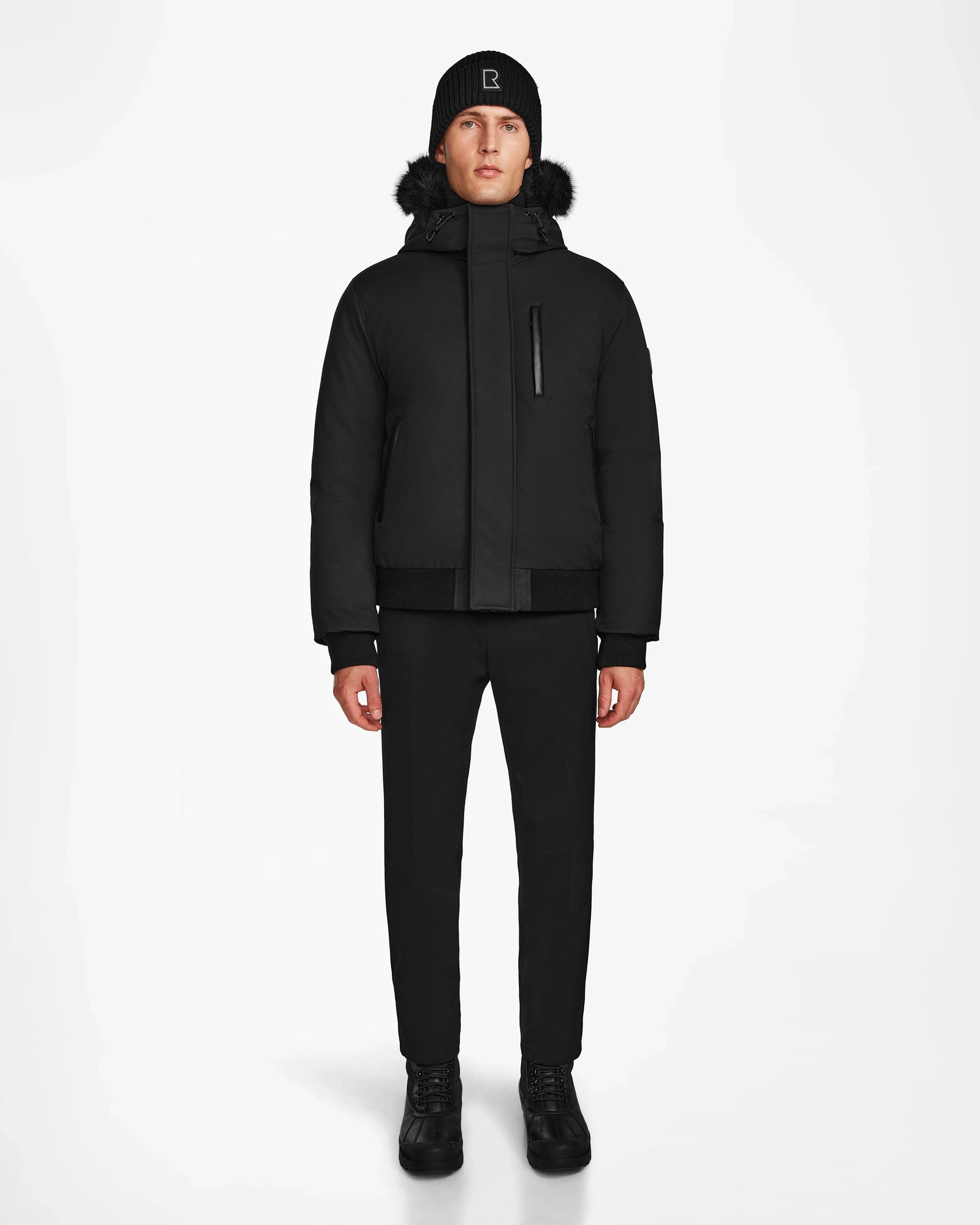 A person faces forward against a white background, wearing the Rudsak DEACON - M123513 Black/Black mens winter coat with a fur-lined hood, paired with black pants, shoes, and a beanie.