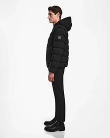 A man stands in profile against a white background, wearing the Rudsak DEMIAN M-R - M123504 Black men’s down puffer made from recycled matte fabric, paired with black pants and boots.