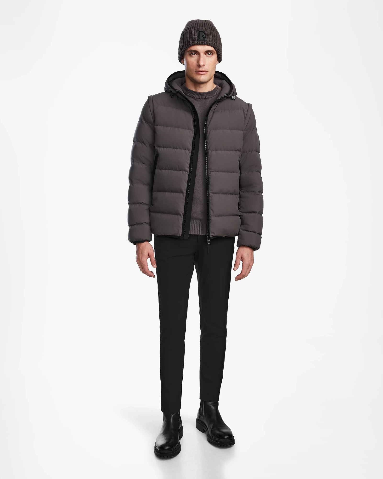 A man faces forward with a neutral expression, wearing the Rudsak DEMIAN M-R - M123504 Grey mens down puffer jacket, paired with dark pants, black boots, a gray knit beanie, and a gray shirt against a white background.