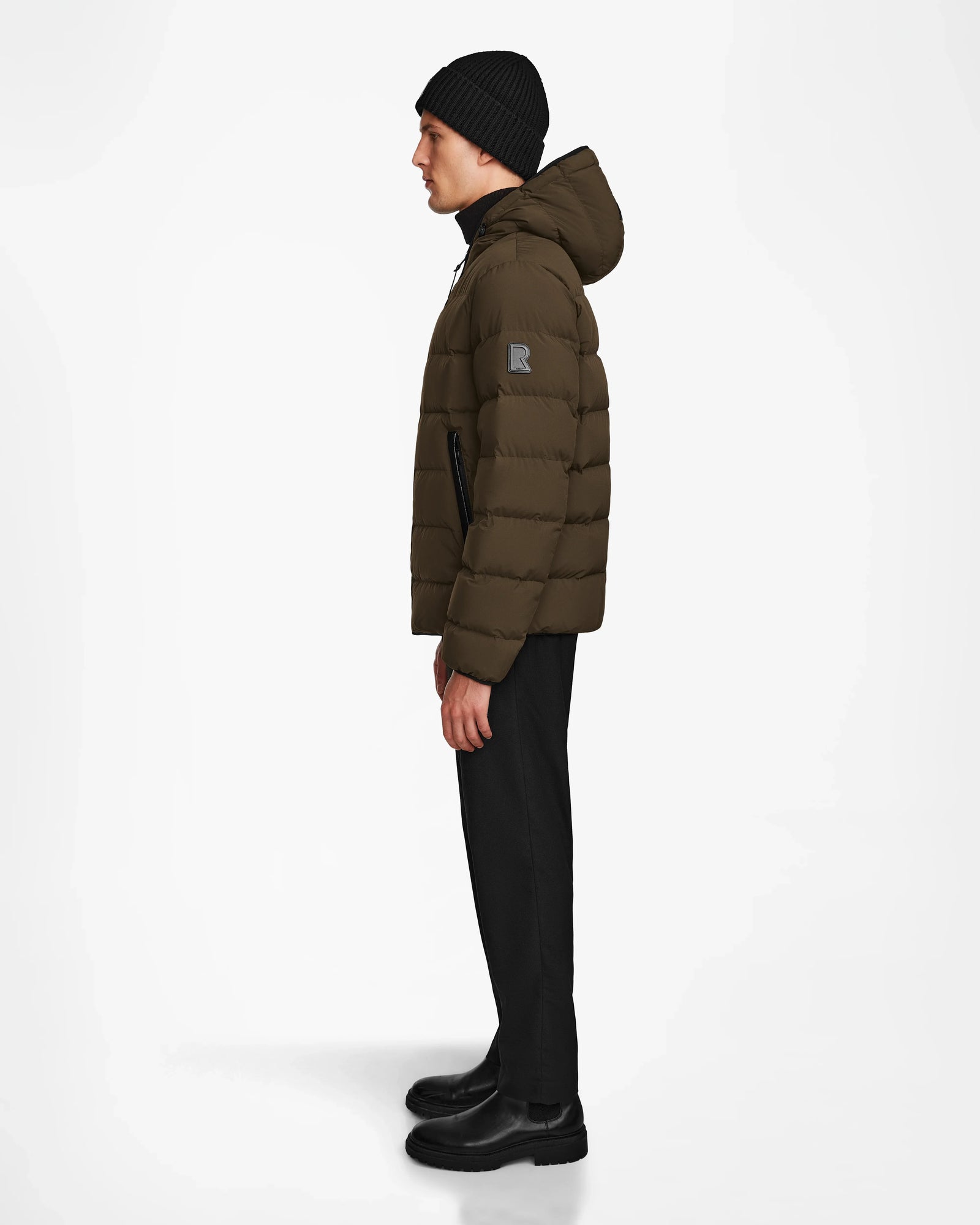 A man wears the Rudsak DEMIAN M - M123507 Army mens down puffer jacket, styled with black pants, a black beanie, and black shoes, standing in profile against a plain white background.