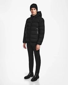 A man stands against a white background, wearing Rudsaks DEMIAN M - M123507 Black hooded down puffer crafted from sustainable recycled fabric, paired with a black beanie, pants, and boots.