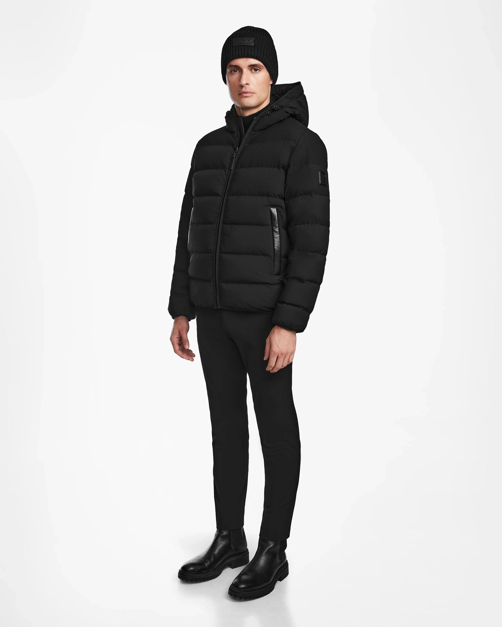 A man stands against a white background, wearing Rudsaks DEMIAN M - M123507 Black hooded down puffer crafted from sustainable recycled fabric, paired with a black beanie, pants, and boots.